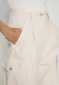 Beige cargobyxor med hög midja, plisserad front, sidfickor och detaljer i silverfärgad metall. Tillverkade av lätt bomullstyg.