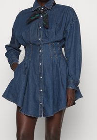 Abito chemisier in denim con chiusura a bottoni frontale, taschino sul petto, vita cin cin e orlo svasato. Presenta maniche lunghe e un accessorio sciarpa nera.