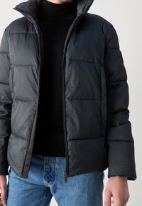Giacca puffa trapuntata nera con colletto alto e chiusura a zip, con un sottile motivo a scacchi sul tessuto. Abbinata a un dolcevita nero.