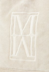 Tissu en éponge beige présentant un logo brodé avec les lettres "M" et "N" dans un cadre rectangulaire. Texture douce avec des détails en relief.