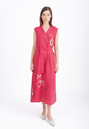 Kobieta w różowej sukience midi bez rękawów z kwiatowym haftem i materiałowym paskiem, w białych butach z ostrym noskiem, stojąca na białym tle.