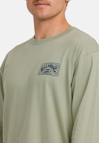 Uomo che indossa un maglione verde chiaro con un piccolo logo Billabong e una grafica tropicale sul lato sinistro del petto.
