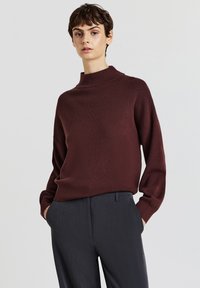 Maglione dolcevita color bordeaux in tessuto morbido e strutturato con vestibilità rilassata e spalle scese, abbinato a pantaloni scuri eleganti.