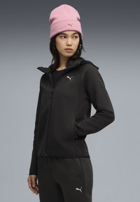 Veste noire zippée avec capuche, poches latérales et accents texturés. Portée avec un bonnet rose et un haut noir, arborant un logo blanc.
