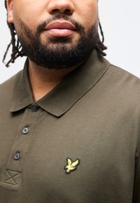 Άνδρας με γενειάδα που φοράει μπλουζάκι πόλο ελιάς με κίτρινο λογότυπο πουλιού στο στήθος, εμφανιζόμενος από τον λαιμό μέχ ρι τη μέση του κορμού.