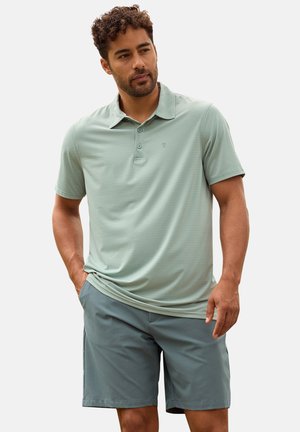 Hombre con cabello rizado que lleva una camisa tipo polo de manga corta color verde claro y pantalones cortos grises, de pie con una mano en el bolsillo.