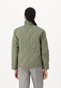 Quiltad olivgrön jacka med hög krage och långa ärmar, prydd med diamantmönster och en lätt textur, i kombination med rutiga byxor.