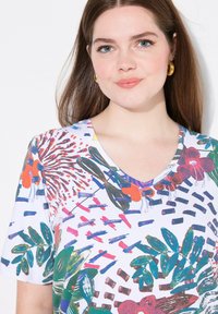Joven mujer con cabello largo castaño que lleva pendientes de aro dorados y una camiseta blanca de cuello en V con patrones florales y abstractos coloridos.