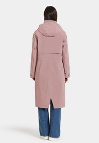 Ljusrosa trenchcoat med avtagbar huva, långa ärmar och en detalj med vent bak, tillsammans med vida jeans i denim och vita skor.