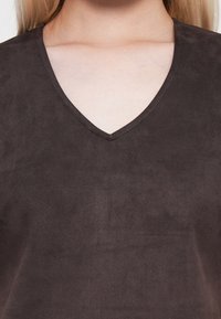 Gros plan d'une personne portant un haut en daim marron foncé à col en V, mettant l'accent sur sa clavicule et la partie supérieure de la poitrine.