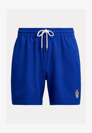 Pantaloni scurți de baie albaștri cu șnur alb și un mic urs brodat care poartă o jachetă, pantaloni și adidași pe partea inferioară a piciorului drept.