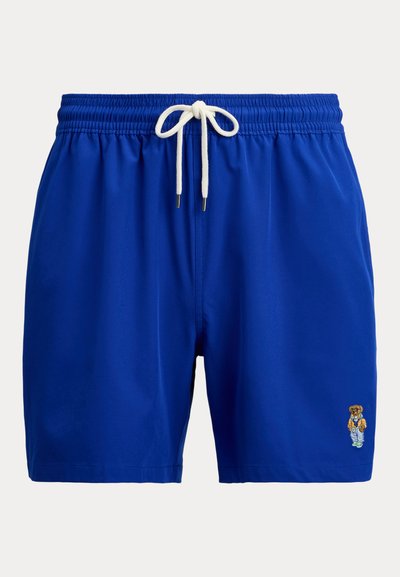 Μπλε βερμούδα για κολύμβηση με λευκό κορδόνι και μικρή κεντημένη αρκούδα που φοράει μπουφάν, παντελόνι και αθλητικά παπούτσια στο κάτω δεξί πόδι.