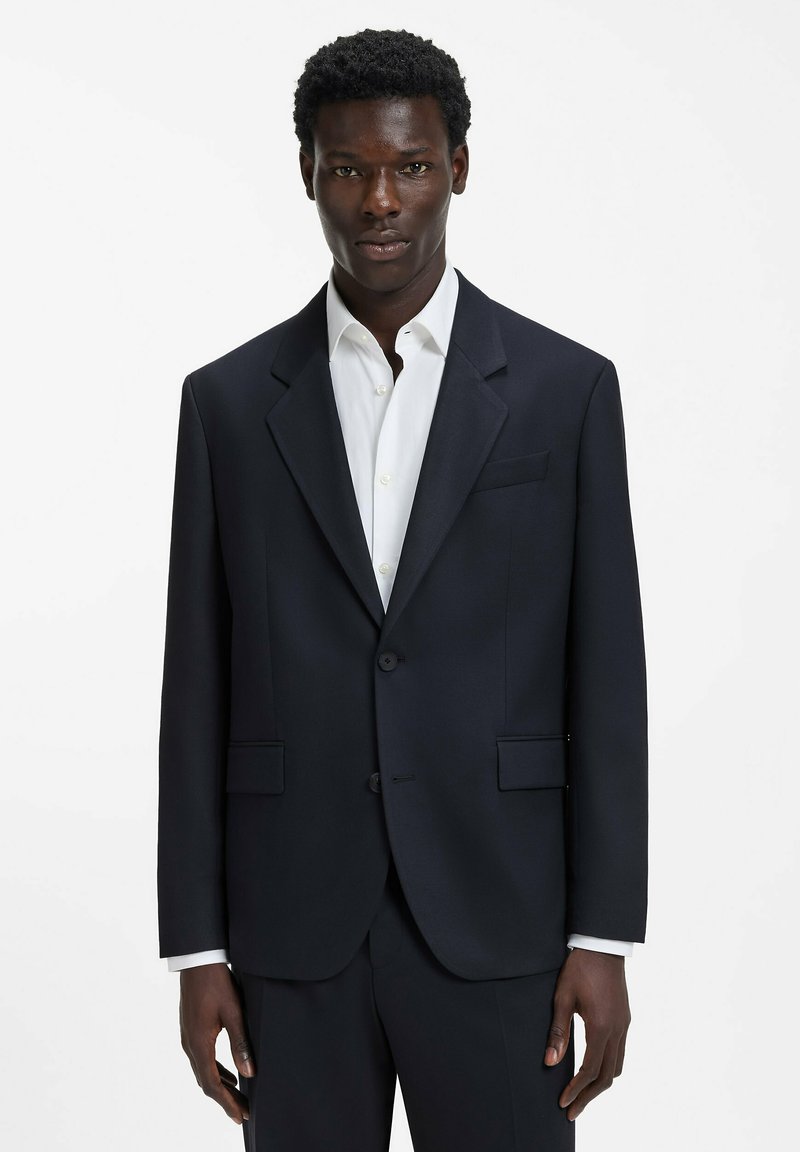 Granatowa marynarka z gładkiej tkaniny, z klapą z wcięciem, dwoma guzikami i bocznymi kieszeniami, zestawiona z białą koszulą pod spodem.