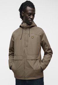 Άνδρας με σκοτεινά πλεγμένα μαλλιά που φοράει καφέ μπουφάν με κουκούλα και κίτρινο λογότυπο πουλιού, με τα χέρια στις τσέπες, κοιτάζοντας προς τα κάτω στο πλάι.