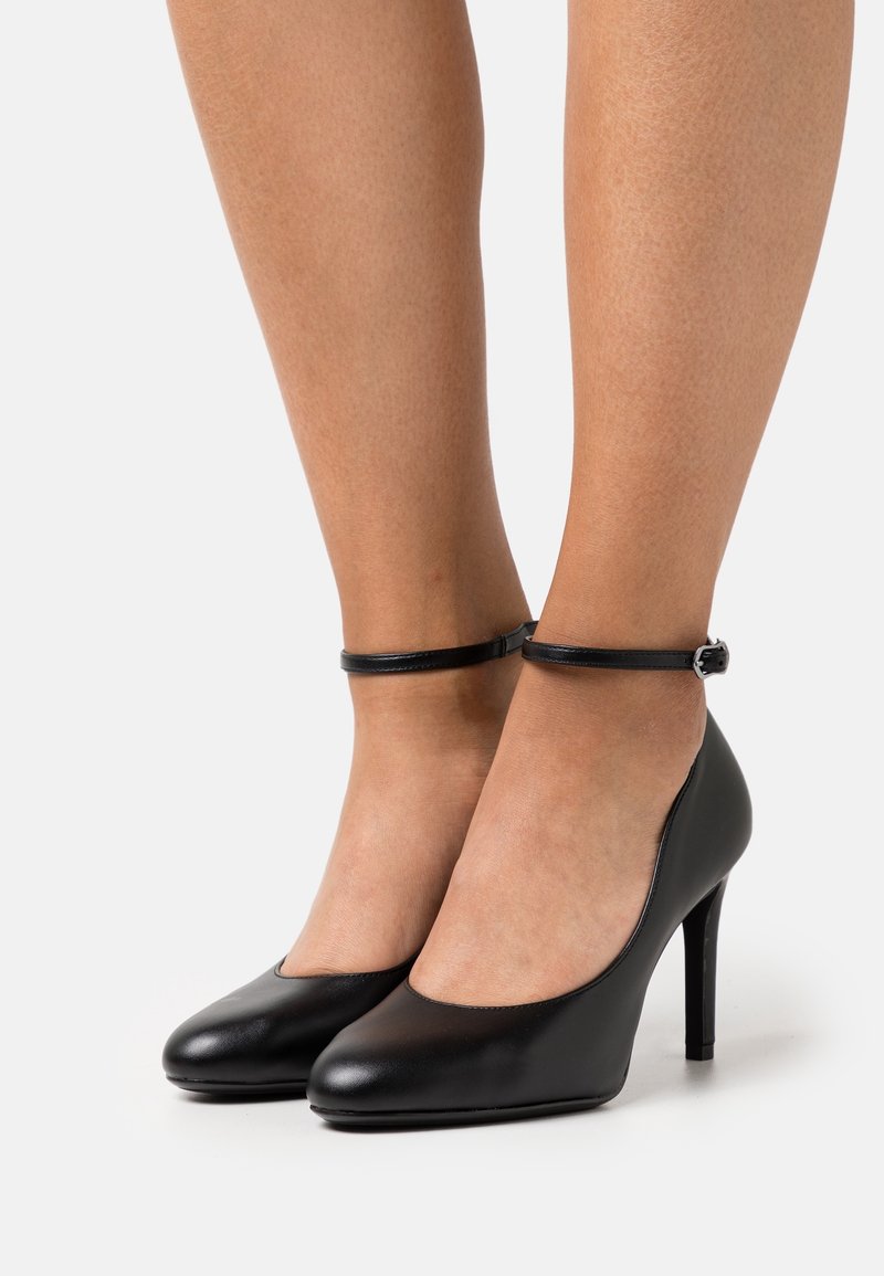 Jambes portant des chaussures à talons hauts en cuir noir avec des brides à la cheville et de petites boucles argentées sur fond blanc.