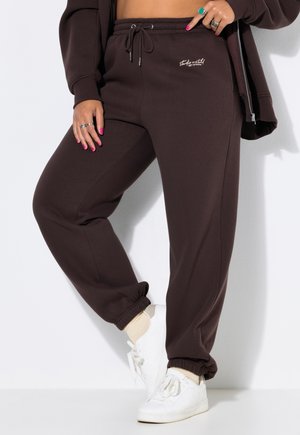 Personne portant un pantalon de jogging marron foncé avec le texte "Studio Untold", des baskets blanches, des chaussettes pâles et une veste foncée, debout devant un fond blanc.