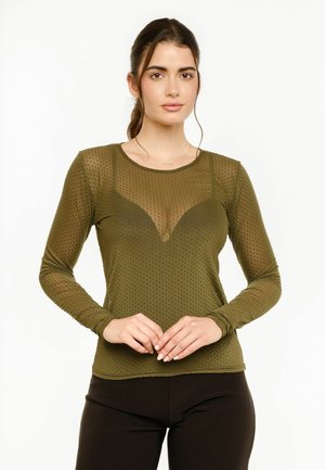 Jeune femme aux cheveux foncés attachés porte un haut transparent vert olive à pois à manches longues sur un bralette assorti, debout les mains jointes.