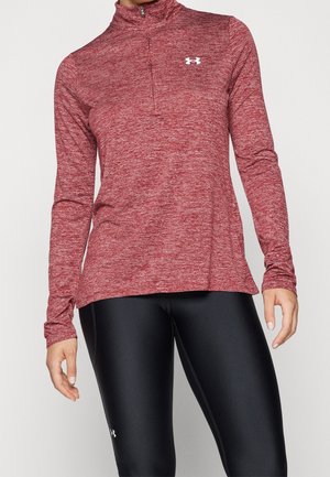 Kobieta ubrana w czerwoną sportową bluzę z długim rękawem i zamkiem do ćwiartek oraz czarne dopasowane legginsy, stojąca na jednolitym tle.