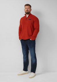 Roter Pullover mit Reißverschluss und strukturiertem Stoff, kombiniert mit dunkelblauen Jeans und weißen Sneakers. Minimalistisches Design mit einem Logodetail.