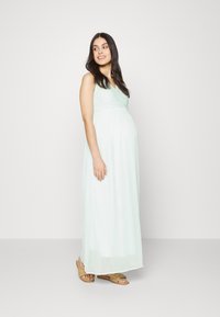 Vestido de maternidade verde menta claro com decote em V, bodice de renda e saia fluida até ao joelho. Usado com sandálias bege. Textura de tecido suave.