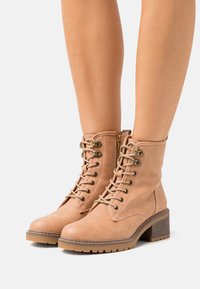 Bottines beige avec une texture lisse semblable au cuir, bout arrondi, lacets à l'avant, accessoires dorés et talon moyen empilé.