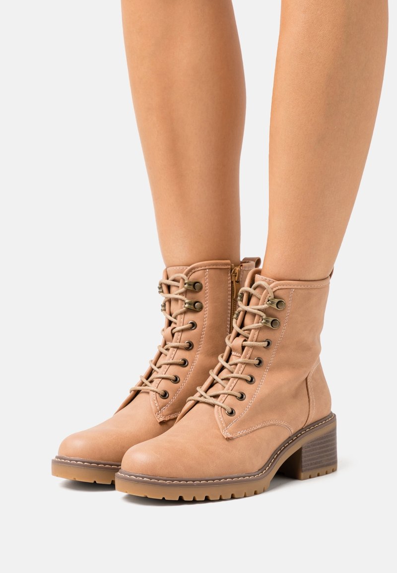 Bottines beige avec une texture lisse semblable au cuir, bout arrondi, lacets à l'avant, accessoires dorés et talon moyen empilé.