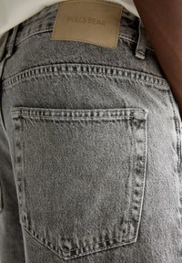 Acercamiento de una persona que lleva vaqueros de mezclilla grises con un bolsillo trasero y una etiqueta de cuero marrón de la marca Pull&Bear encima del bolsillo.