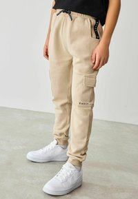 Beige kravas bikses, kas izgatavotas no mīksta materiāla, ar elastīgu jostasvietu, divām sānu kabatām un drukātu zīmola logo uz kājas. Apvienotas ar baltām sporta apavām.