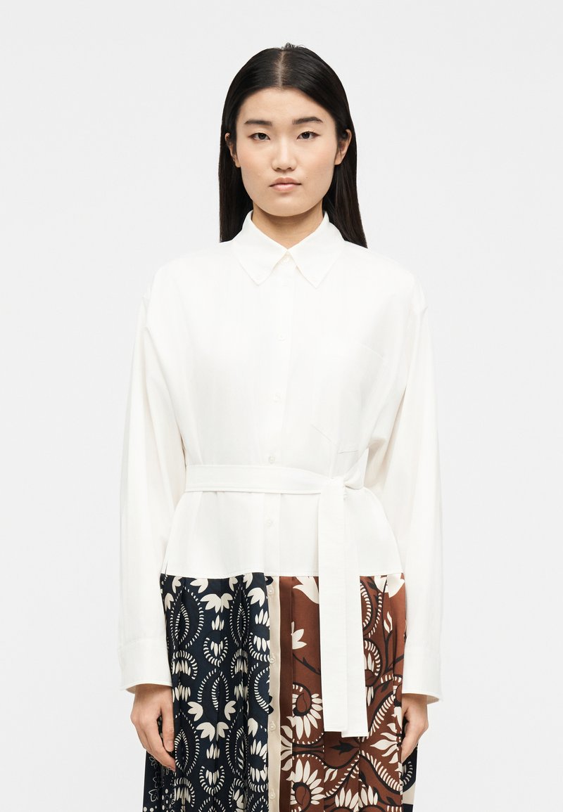 Γυναίκα με ίσια μαύρα μαλλιά φοράει λευκό πουκάμισο με κουμπιά και ζώνη, καθώς και φούστα με ναυτικό μπλε και καφέ floral μοτίβα, στέκεται με φόντο το λευκό.