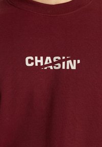Bordo džemperis su apvalia iškirpte, su centrine baltos spalvos grafika, kurioje dalis žodžio "CHASIN‘" kertama perpjautu brūkšniu.