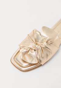 Chausson en métal doré avec un grand nœud décoratif sur le dessus. Présente un bout carré et une surface lisse et texturée.
