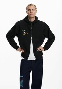 Felpa nera con zip e grande cappuccio, con design ricamato di Topolino. Abbinata a pantaloni in denim scuro con una toppa decorata.