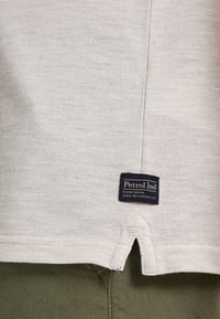 Vit bomullsskjorta med ventilerad nederkant, försett med en mörkblå vävd etikett med texten "Petrol Ind" och synlig textilstruktur.