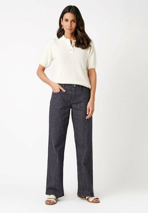 Woman standing with right hand in pocket, wearing white short-sleeve polo shirt, navy pinstriped wide-leg pants, and white sandals.