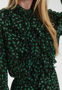 Grön blommönster med svart bakgrund, med volangdetaljer, hög halsringning och genomskinligt, lätt tyg. Knappar synliga vid kragen.