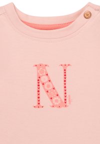 Ljust rosa bomullströja med ett framträdande rött bokstav "N" som har blommönster och hjärtan, rund halsringning och tryckknappdetalj.