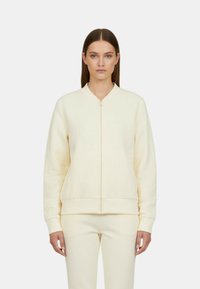 Felpa crema con zip e lettere in rilievo, polsini e orlo a coste, tessuto leggero e vestibilità ampia con pantaloni coordinati.