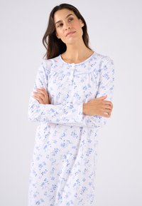 Camisa de noche de manga larga en tejido blanco, con un estampado floral en azul. Cuello redondo con cierre de botón y detalles fruncidos.
