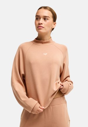 Junge Frau mit hellbraunen Haaren in einem niedrigen Dutt, trägt einen lockeren beigen Pullover und passende Hosen, schaut leicht zur Seite.