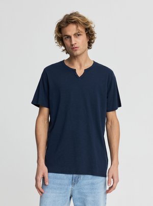 Giovane uomo con capelli biondi ricci che indossa una maglietta henley blu navy a maniche corte e jeans azzurri, in piedi davanti a uno sfondo uniforme.