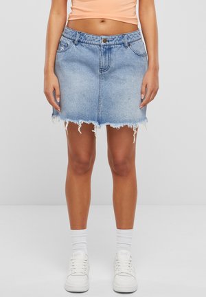 Jupe en denim bleu clair avec une coupe ajustée, ourlet effiloché, cinq poches et un système de fermeture à bouton à la taille. Associée à des baskets blanches et des chaussettes.