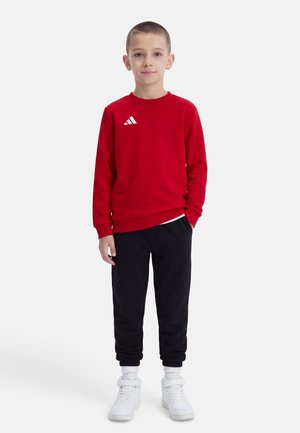 Ragazzo in piedi che indossa una felpa rossa, pantaloni jogger neri, sneakers alte bianche e calzini bianchi su uno sfondo bianco semplice.