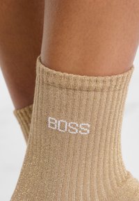 Calcetines dorados con textura suave, que presentan el logo blanco "BOSS" en el puño. El material tiene un ligero brillo y un patrón de tejido consistente.