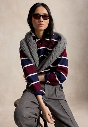 Femme portant des lunettes de soleil, un t-shirt rayé à manches longues, un pull gris posé sur les épaules et un pantalon gris, assise sur un fond neutre.