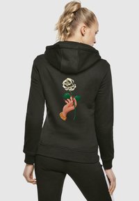 Felpa nera con cappuccio, caratterizzata da una grafica di una mano che tiene una rosa bianca sul retro. Presenta un cappuccio con cordino e polsini e orlo a coste.