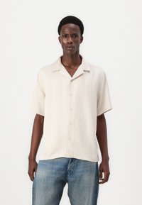 Kortermet, kremfarget skjorte med vertikale striper, åpen krage og fem knapper. Kombinert med lys blå denimjeans.