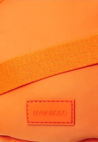 Oransje stoffeveske med en teksturert stropp og en sydd logo-patch. Overflaten er jevn med en ensfarget nyanse og uten synlige mønstre.