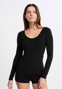 Kobieta stoi przodem, ubrana w dopasowaną czarną bluzkę z długim rękawem i czarne szorty na tle gładkiego białego tła.