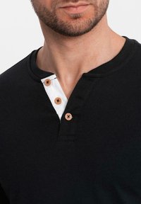 Schwarz-Henley-Shirt mit einer weißen Blende und drei braunen Knöpfen, rundem Halsausschnitt und glatter Baumwolltextur. Nahaufnahme.