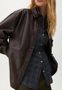 Brun konstläderjacka med tryckknappar, bärs över en mörkgrön rutig skjorta med vita och marinblå toner. Slät textur, avslappnad passform.